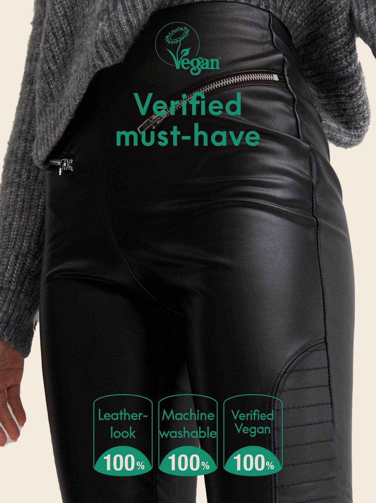 A model walks towards the camera while looking towards the left at a thin golden glitter sculpture, they are wearing a grey v neck cardigan, a pair of black leather look leggings and a pair of heeled ankle boots