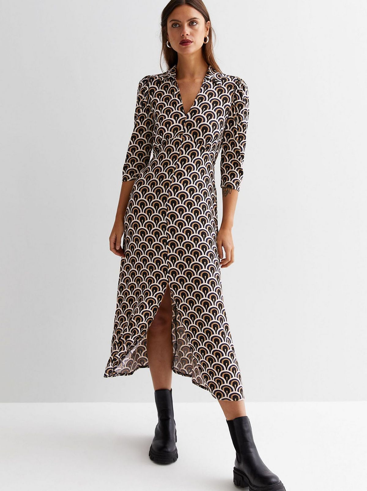 A model stands looking away from the camera in front of a white background, wearing an off white geometric collared midi dress and a pair of black ankle boots