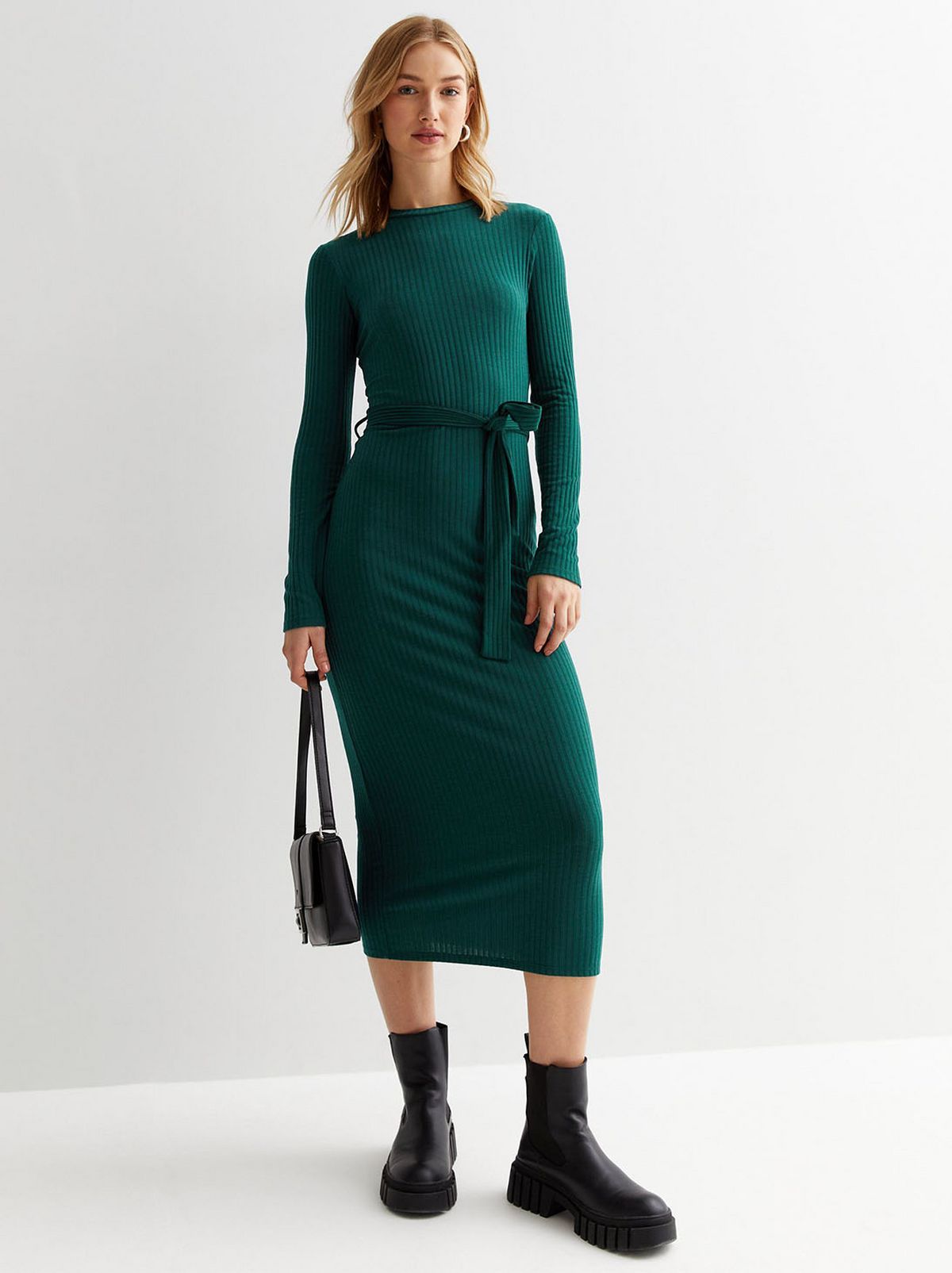 A model stands looking at the camera in front of a white background, wearing a dark green jersey bodycon midi dress and a pair of black ankle boots