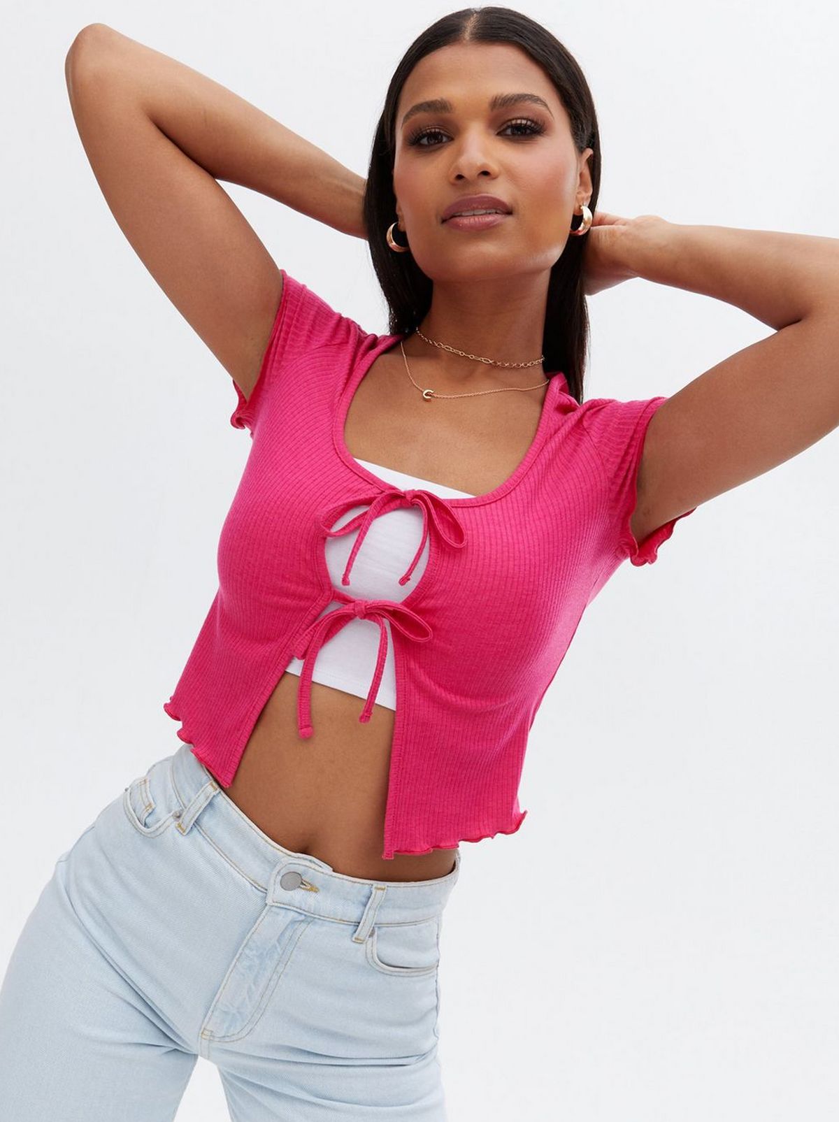 There is a women standing with her hands behind her head staring into the camera wearing a bright pink frill top and a pair of pale jeans