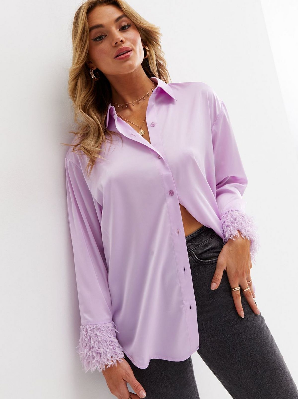 A women leans on a white wall wearing a lilac satin long sleeve shirt and a matching handbag, as well as a pair of grey jeggings