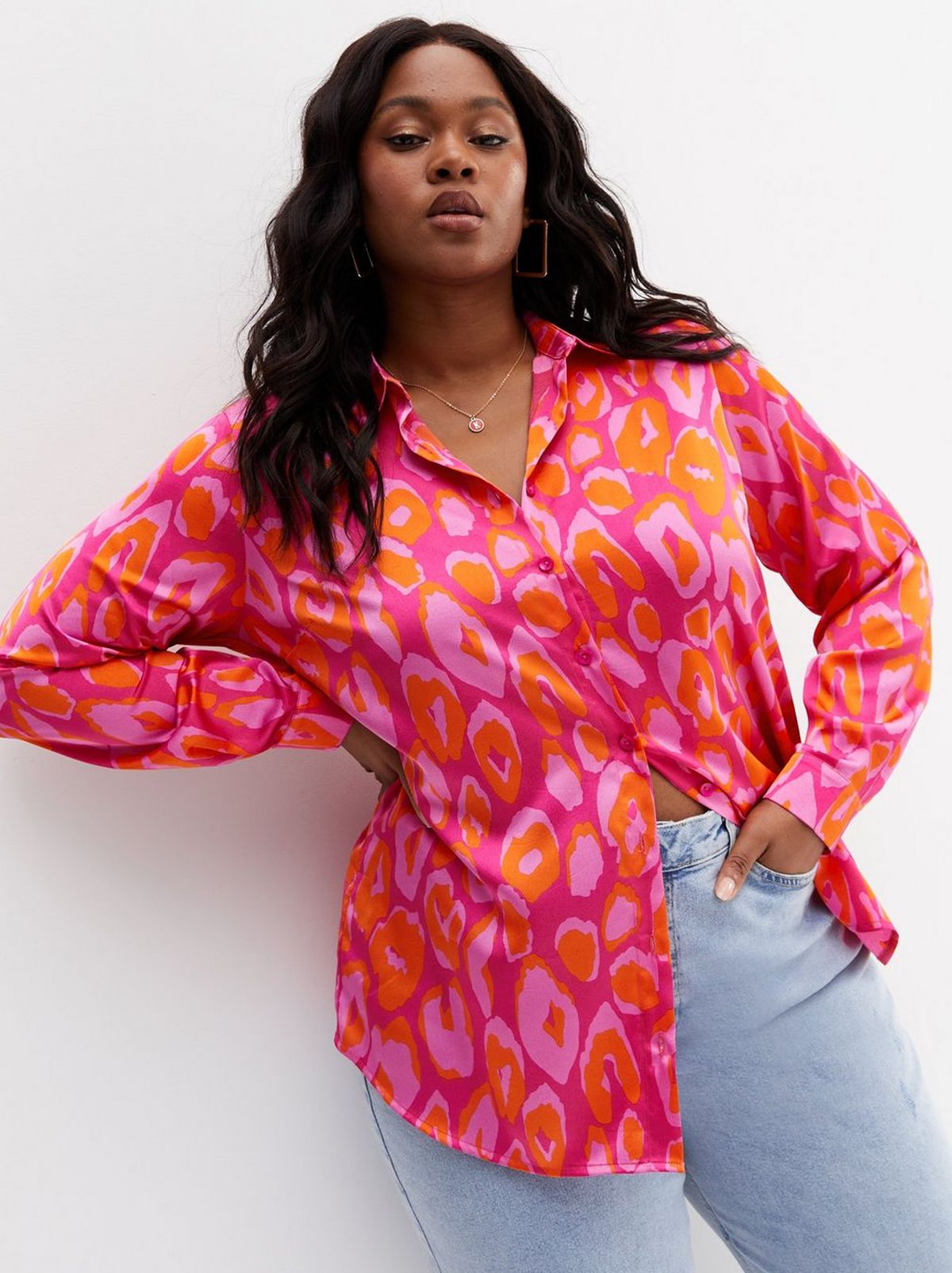 A women wearing an oversized pink satin shirt and a pair of blue denim jeans stares at the camera