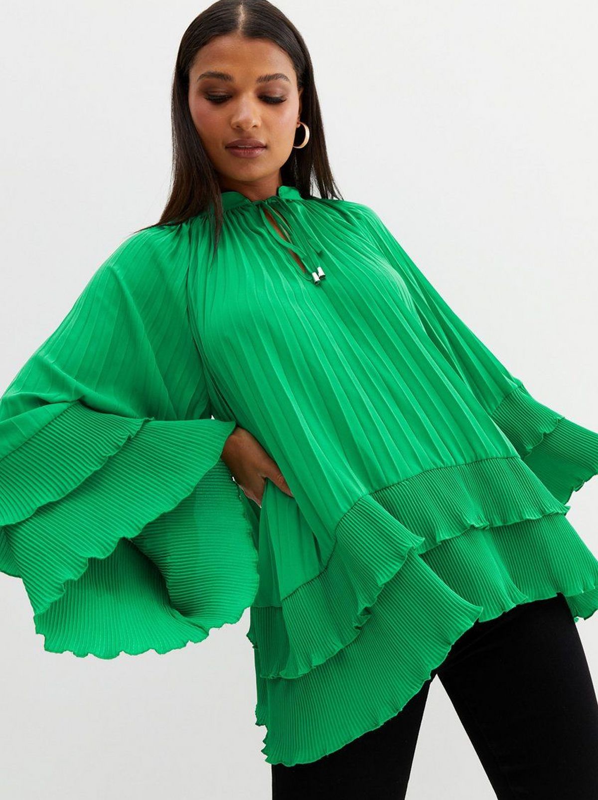 There is a women staring at the floor wearing a green pleated tiered blouse and a pair of black trousers