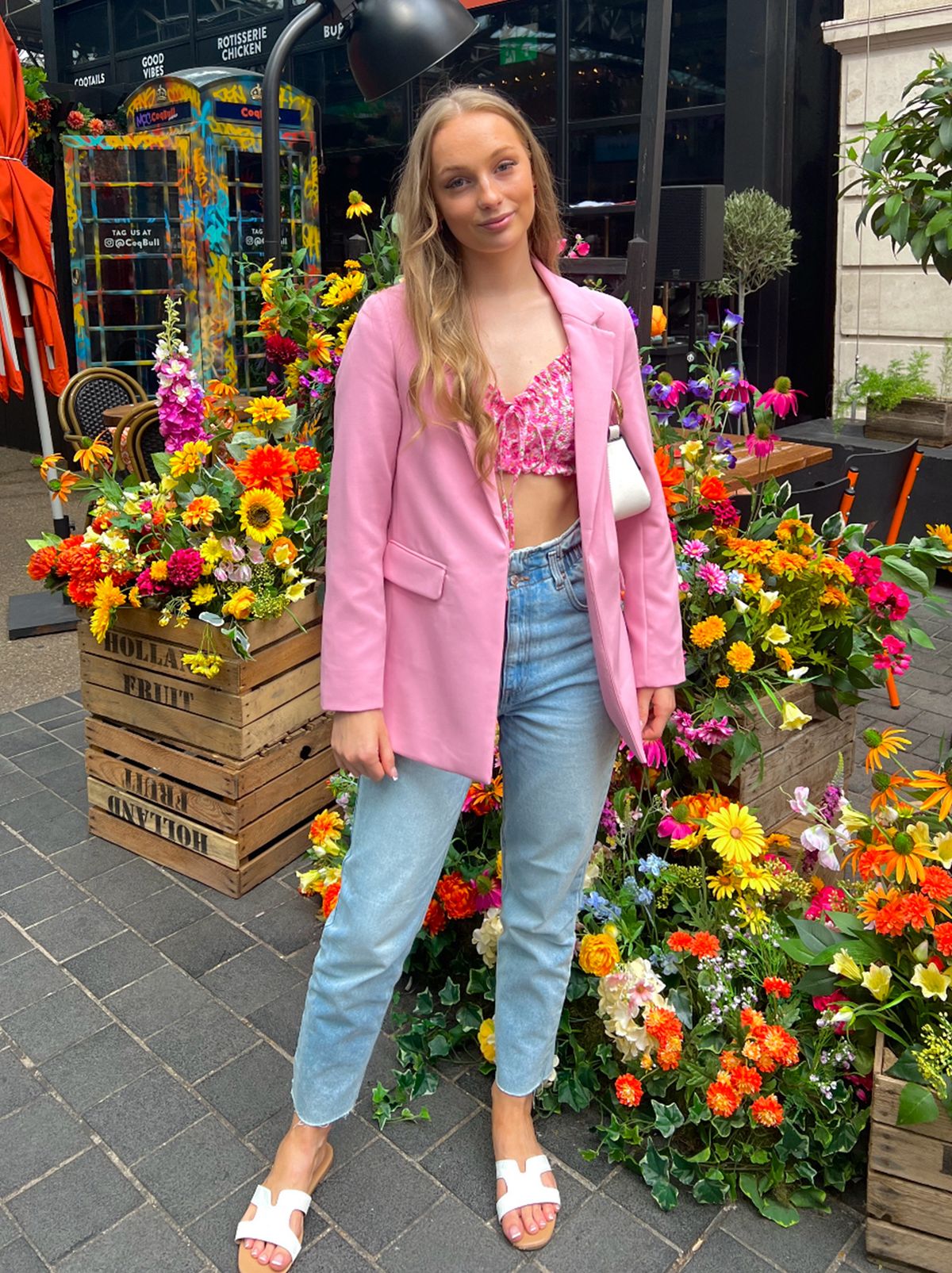 Mabel wears a pink blazer, short pink top and a pair of blue denim jeans standing staring at the camera in front of crates of flowers