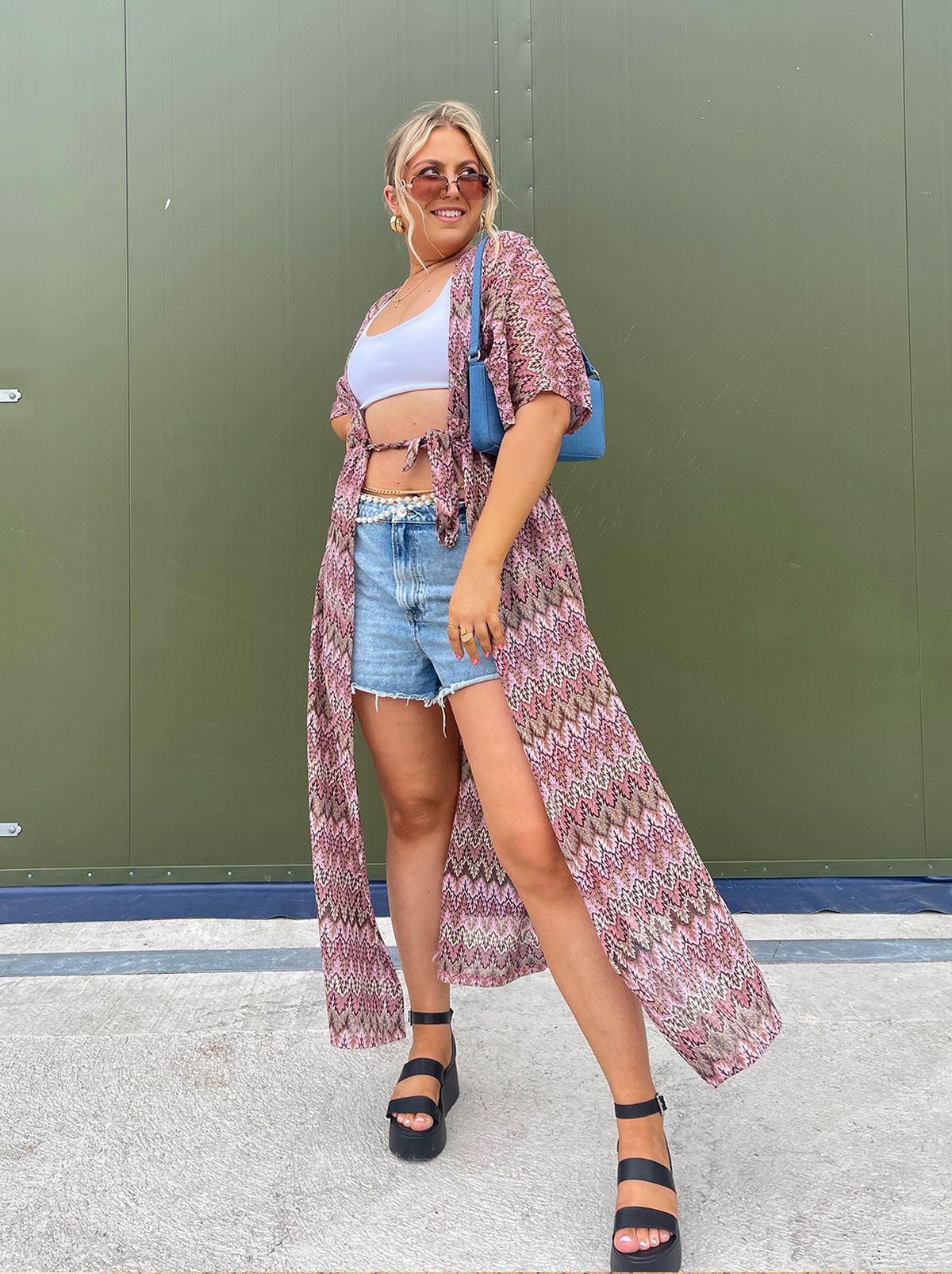 Lauren wears a pair of denim shorts, a short white top and a long summer shawl in front of a green metal door staring at the camera	