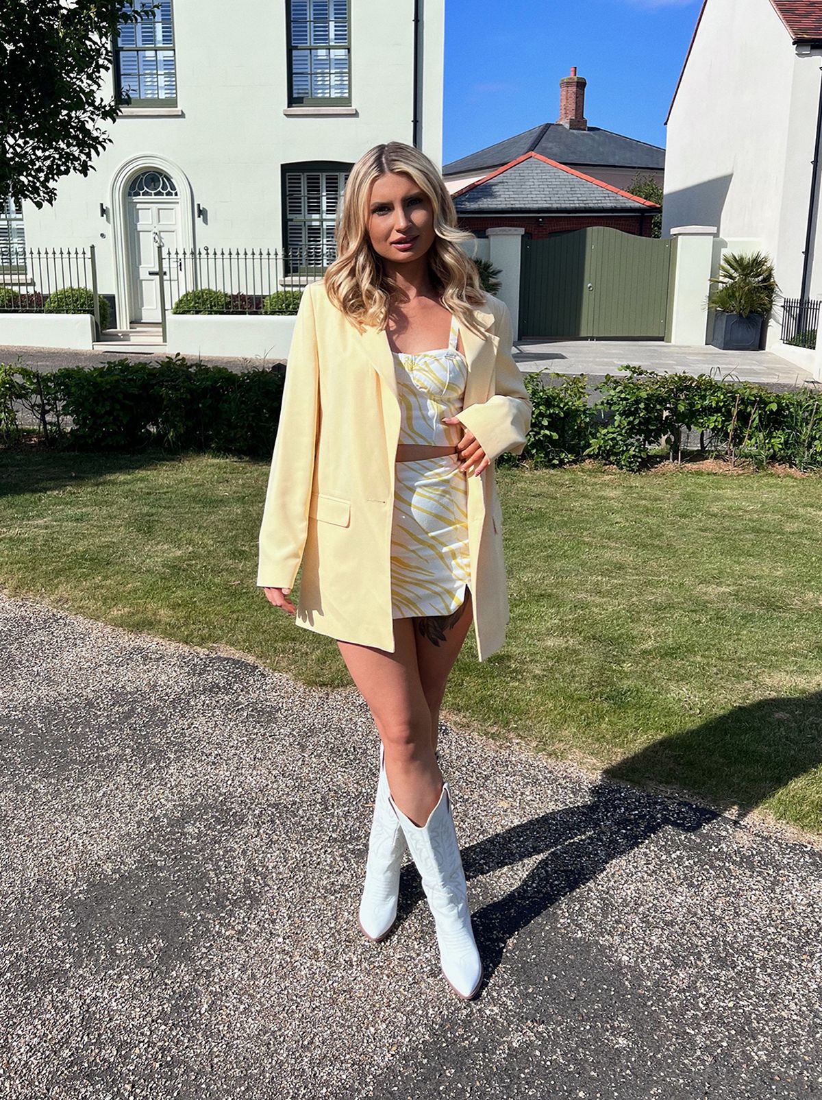 Beth wears a pair of cowboy boots, a light yellow blazer and a matching yellow and white patterned denim skirt and crop top, while standing in a park staring at the camera	