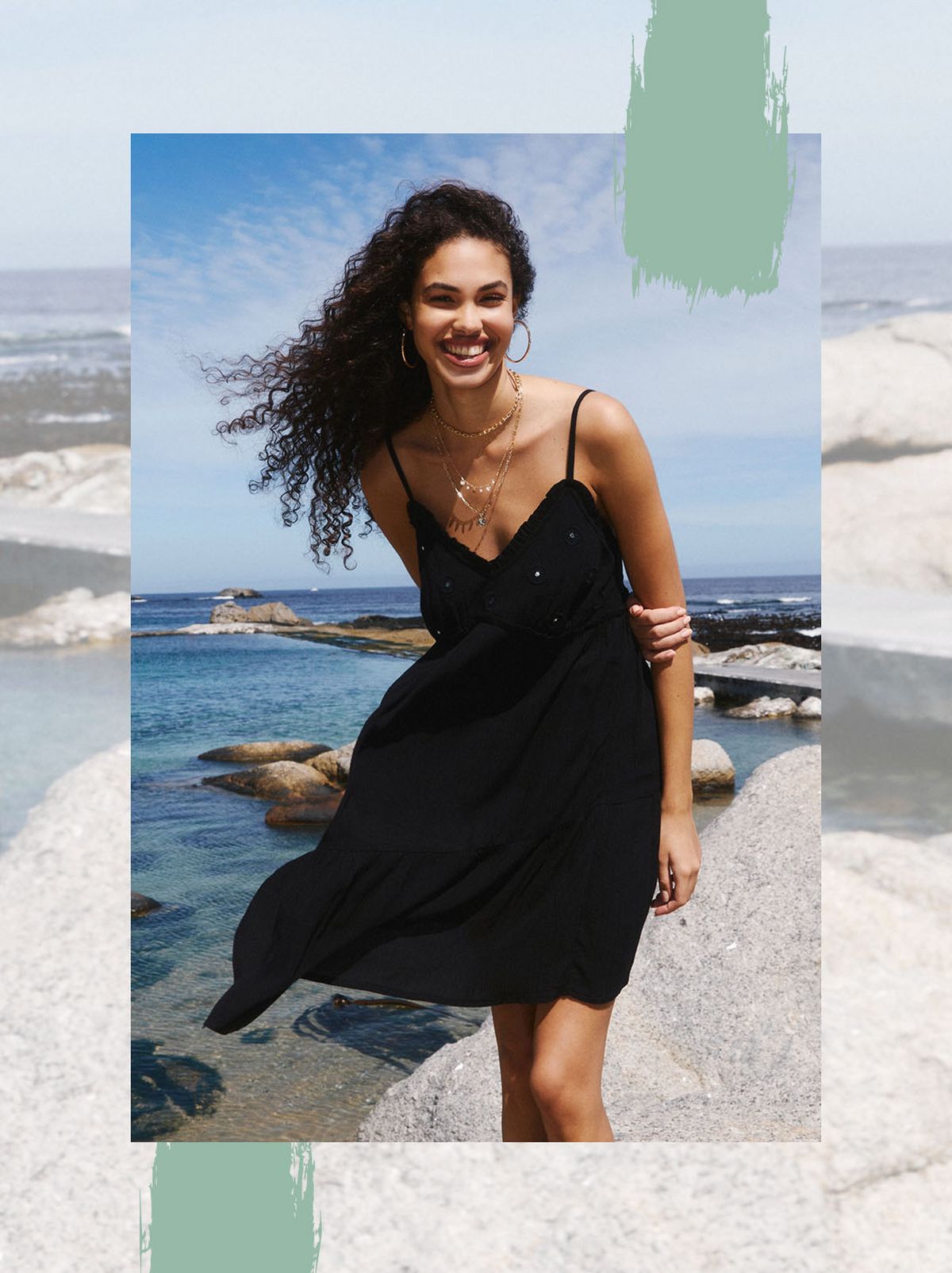 A model stands on a rock at the sea smiling and looking just away from the camera, they are wearing a black mini dress and a pair of black sandals
