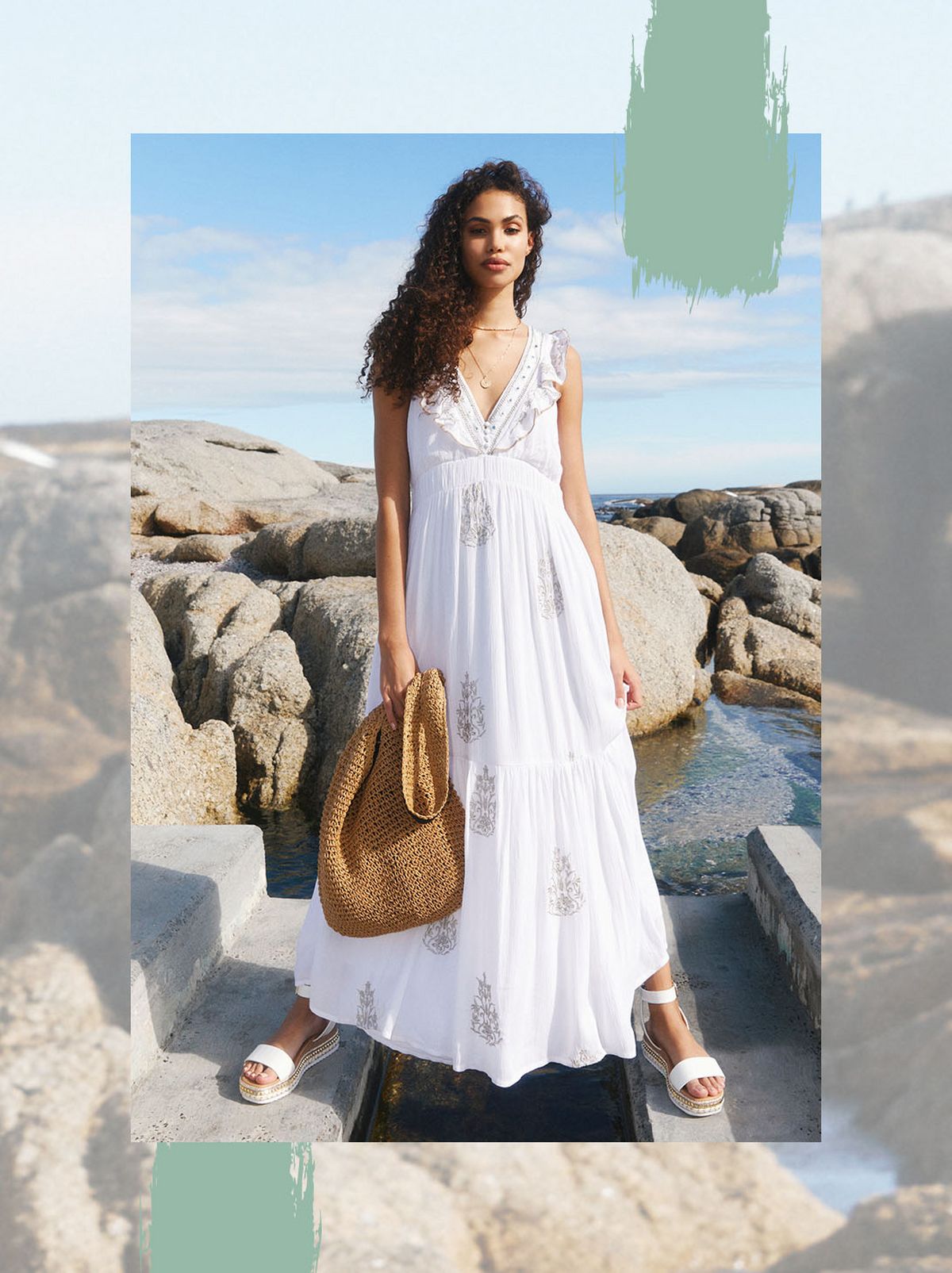 A model stands on a pathway in front of the sea, they are touching their hair and looking away from the camera while wearing a printed dress and pair of black heeled sandals