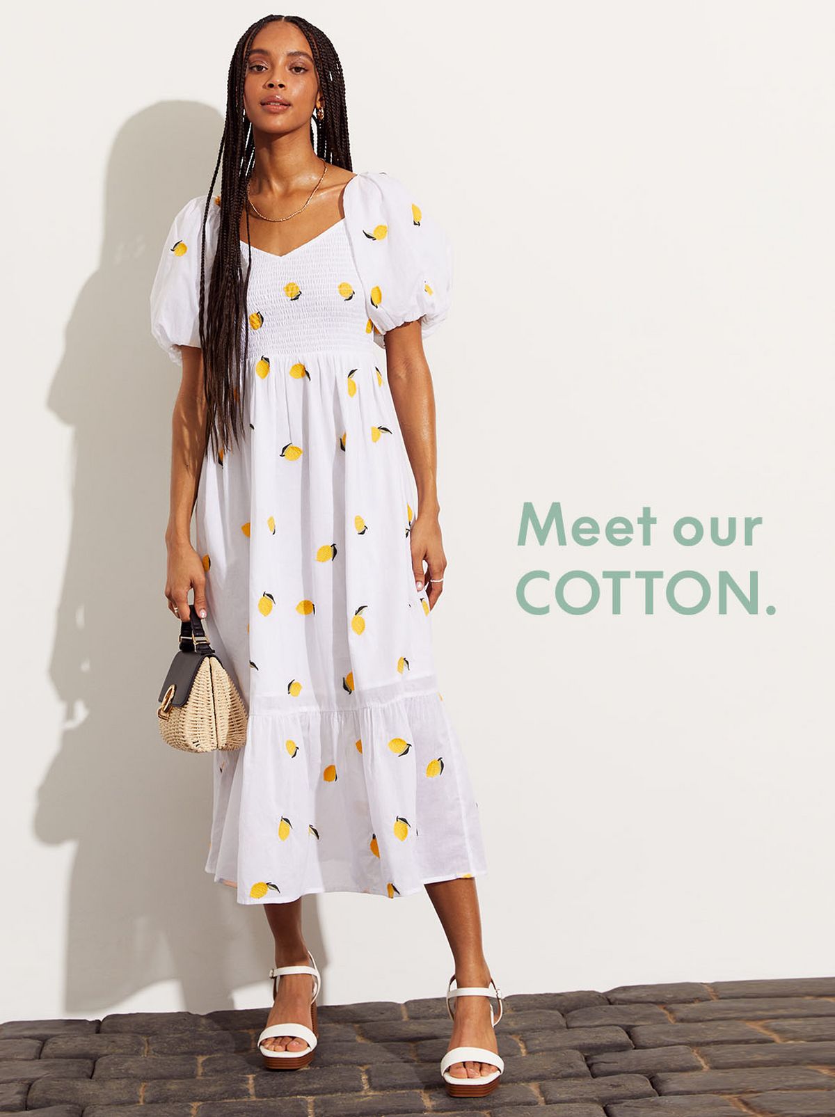 A model stands in front of the camera looking directly at it in front of a white wall while wearing a long cotton dress and a pair of sandals ​