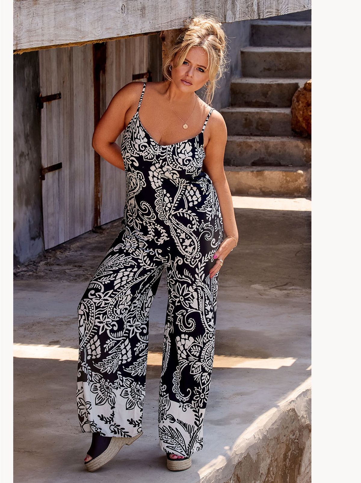 Emily Atack stands with a hand on her hip outside while wearing a black and white patterned jumpsuit​