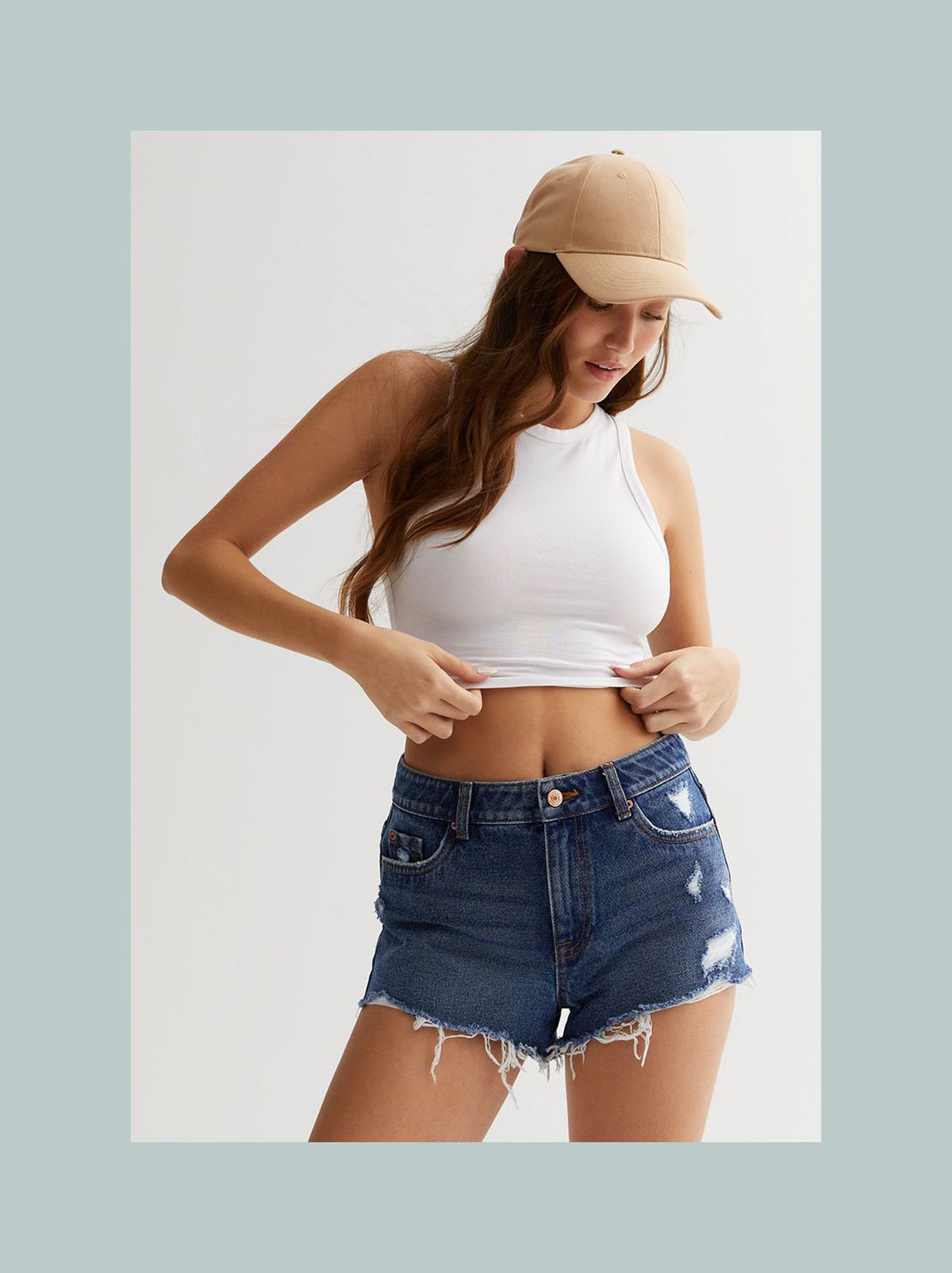 A model is standing in front of a white background wearing a beige cap, a white vest top and a pair of blue denim shorts​