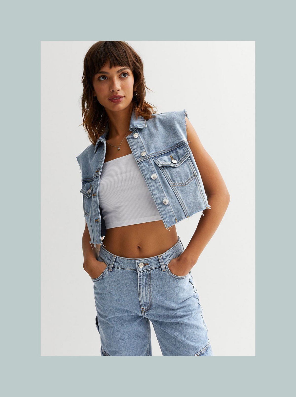 A model is standing in front of a white background wearing a blue denim gilet and a white crop top​