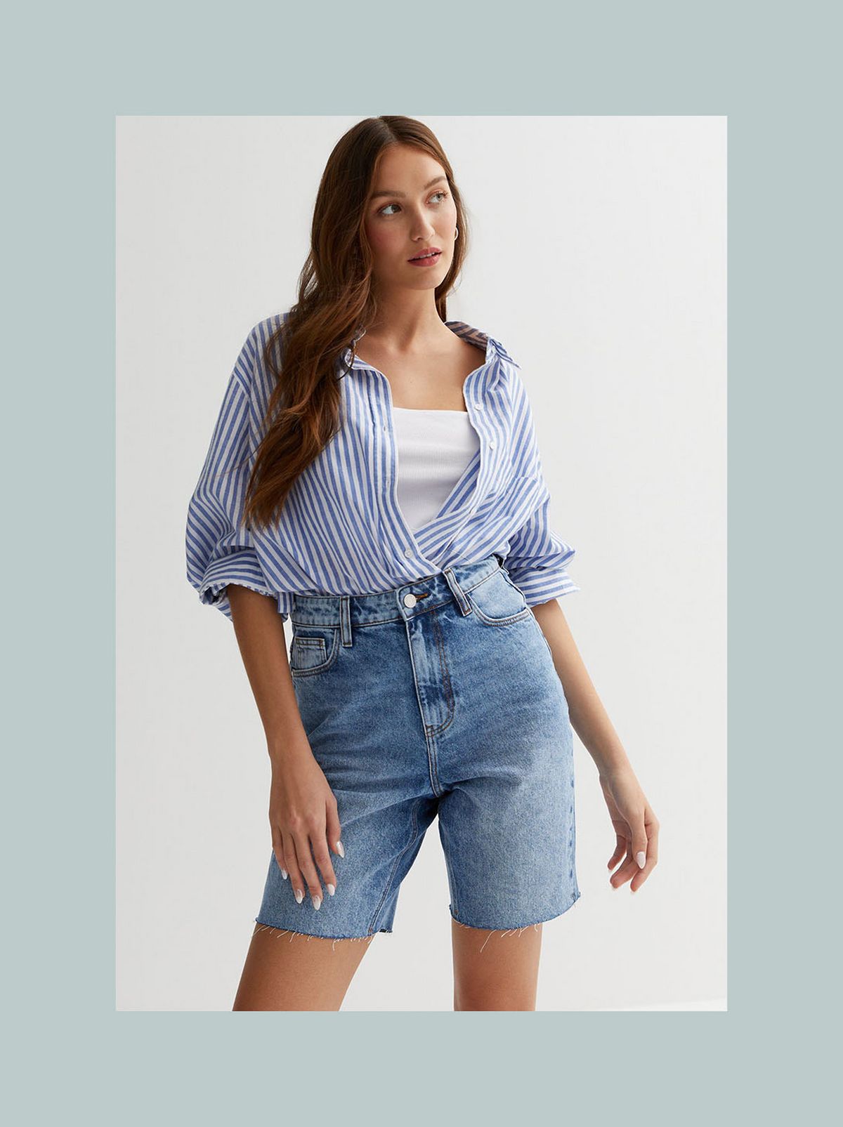 A model is standing in front of a white background wearing a pair of blue high waisted Bermuda shorts, a white top and a striped shirt​