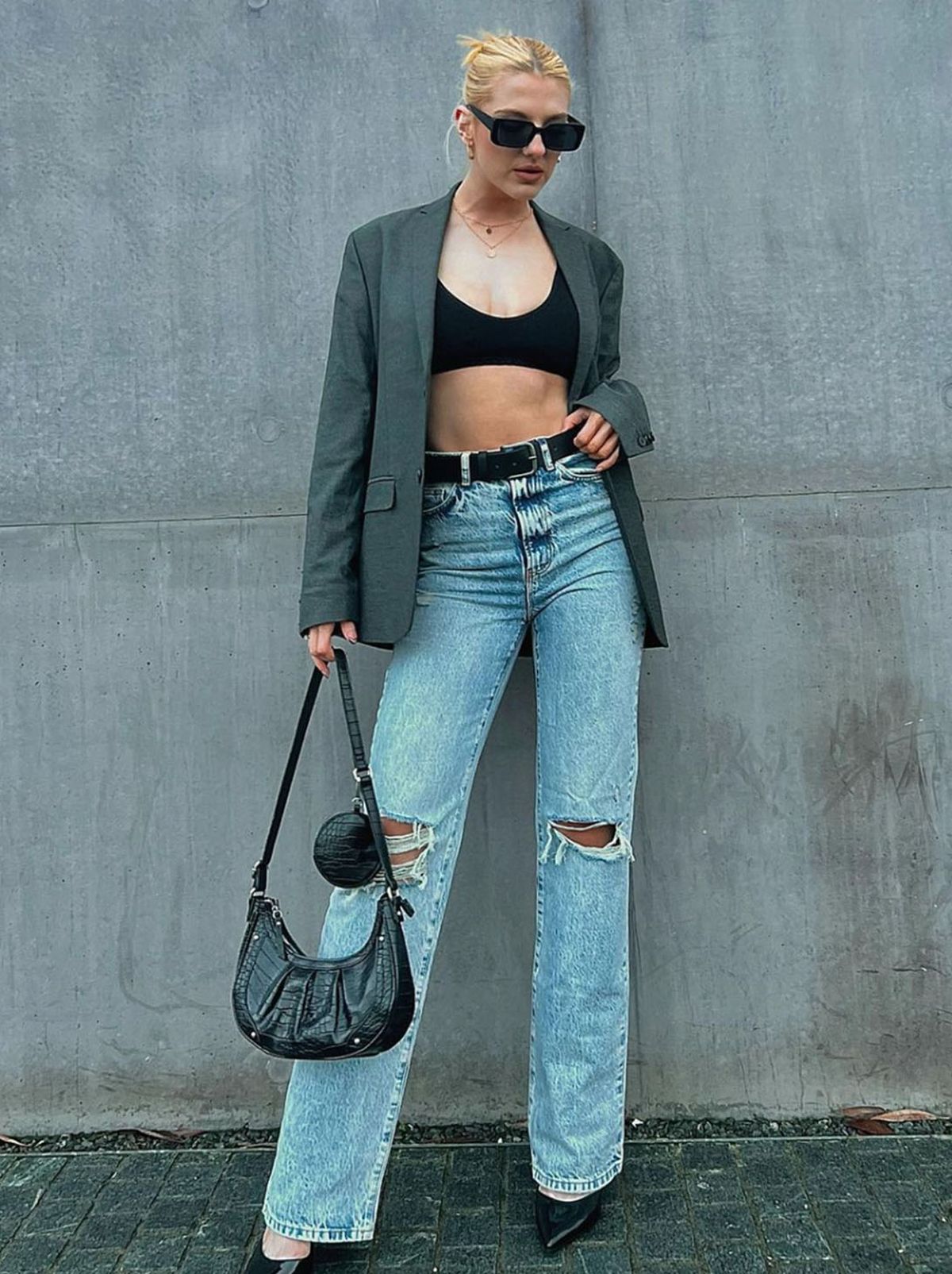 A New Looker stands in front of a blank grey concrete wall looking at the camera while wearing a pair of black sunglasses, a grey blazer, a pair of blue straight leg jeans and a pair of black leather shoes	