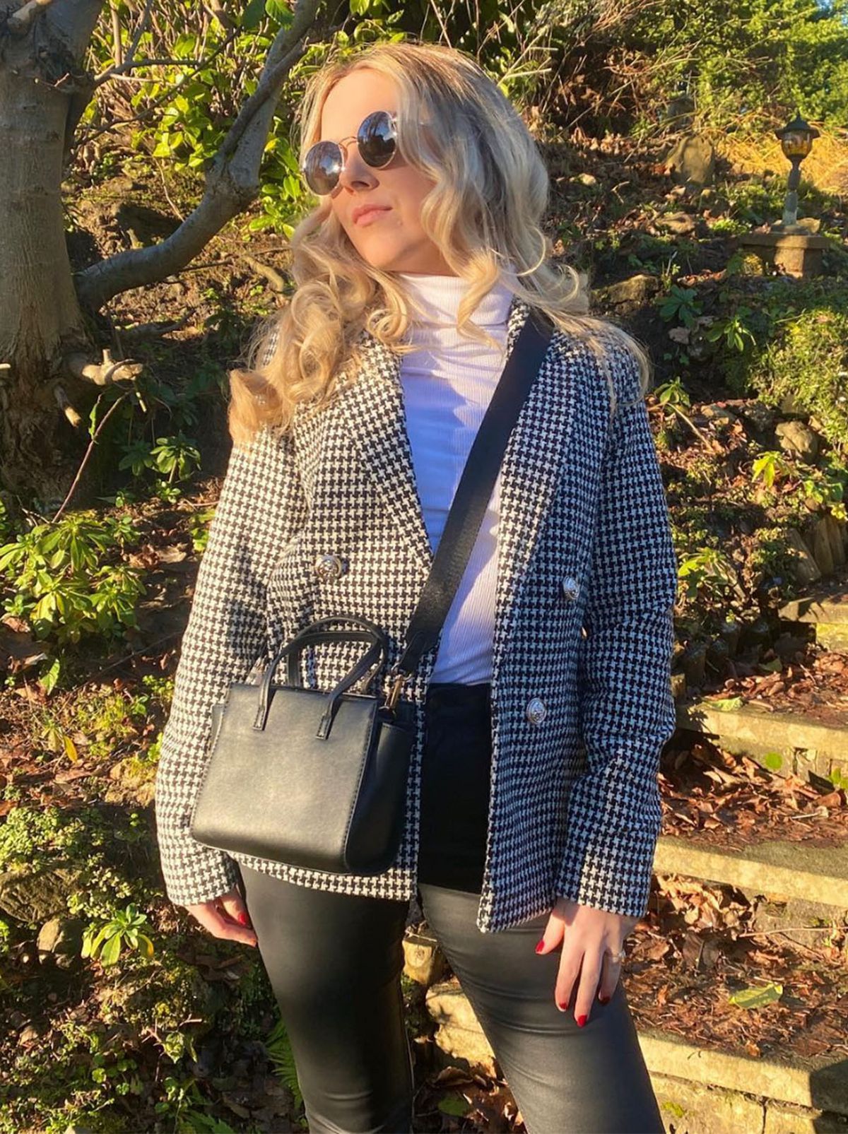 New Looker Caroline is looking away from the camera looking towards the sun while wearing a checkered blazer, a white turtleneck and a black crossbody bag	