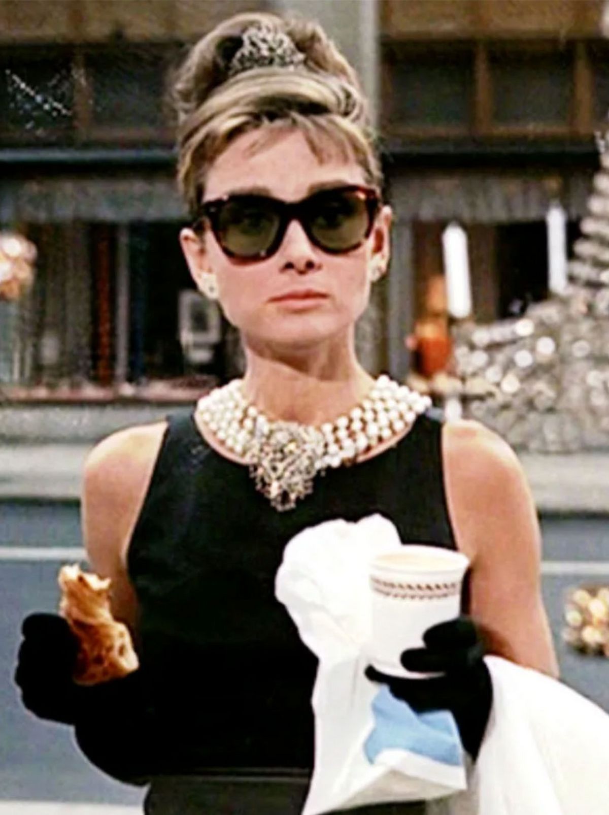 Audrey Hepburn stands on a pavement while looking just next to the camera while holding a pastry, a coffee and a plastic bag