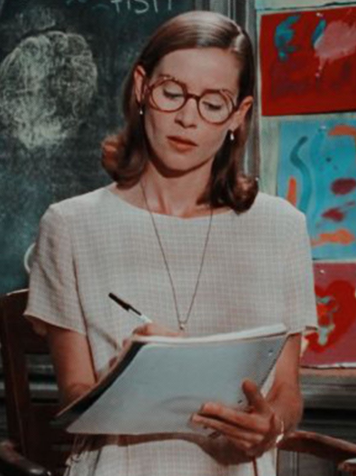 Ms. Wormwood looks down at her book and is writing in it in front of a classroom