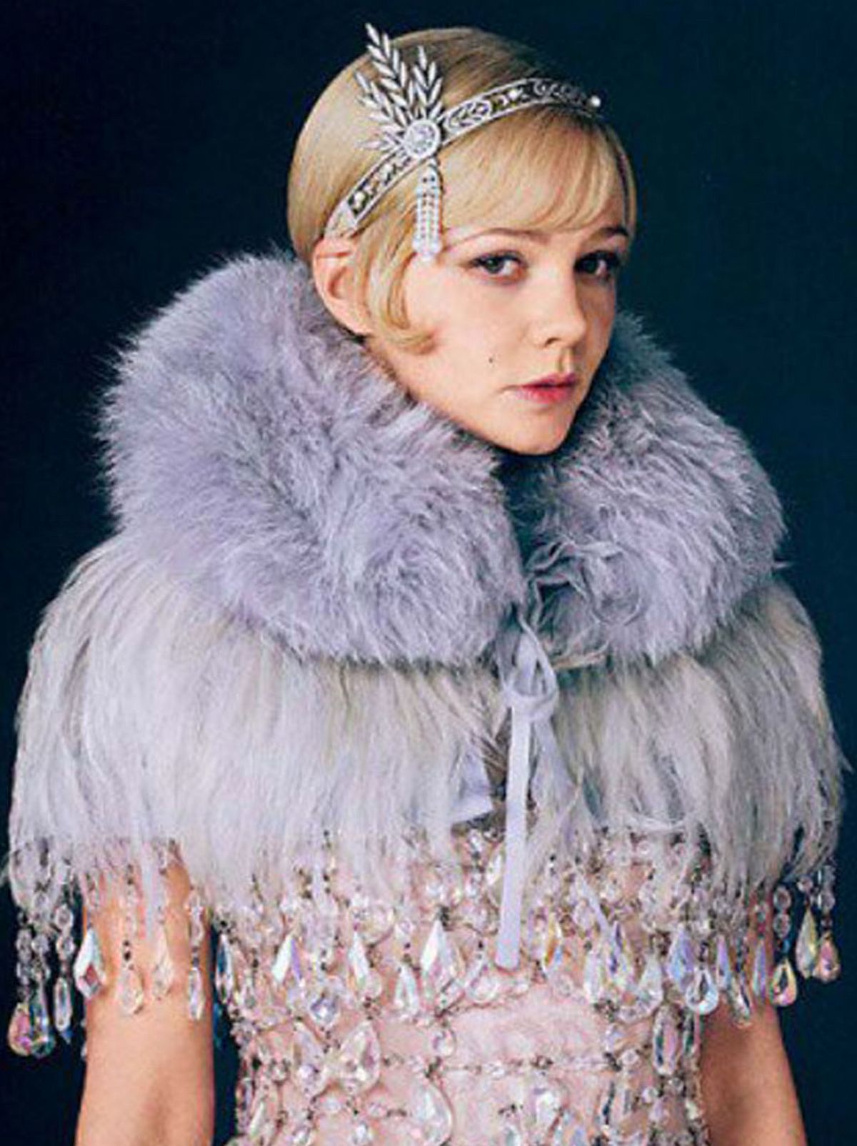 An actress looks at the camera while wearing a 20s era dress, a scarf and a tiara 