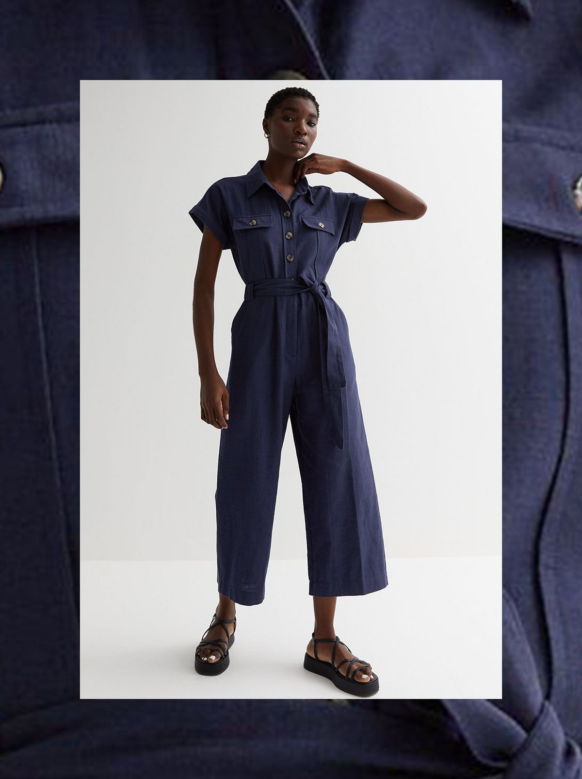 A model stands in front of a white background wearing a navy tie waist short sleeve jumpsuit	
