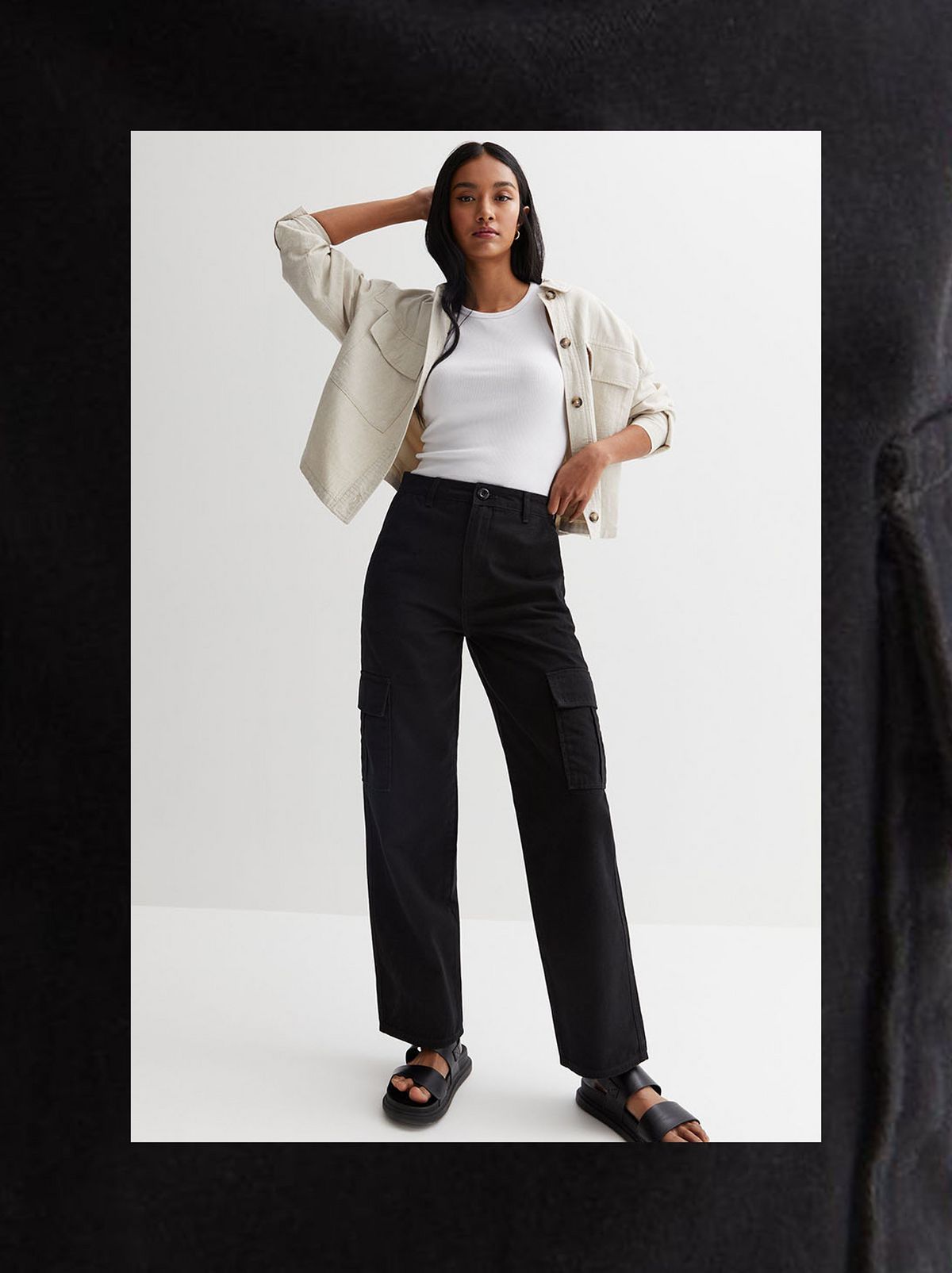 A model stands in front of a white background, touching their hair and looking at the camera while wearing a white vest, a cream overshirt and a pair of black slim fit cargo trousers	
