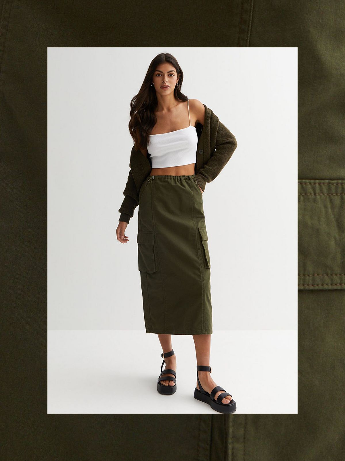 A model stands in front of a white background with a hand in their pocket and wearing a khaki midi skirt, a white vest and a brown zip up hoodie 	