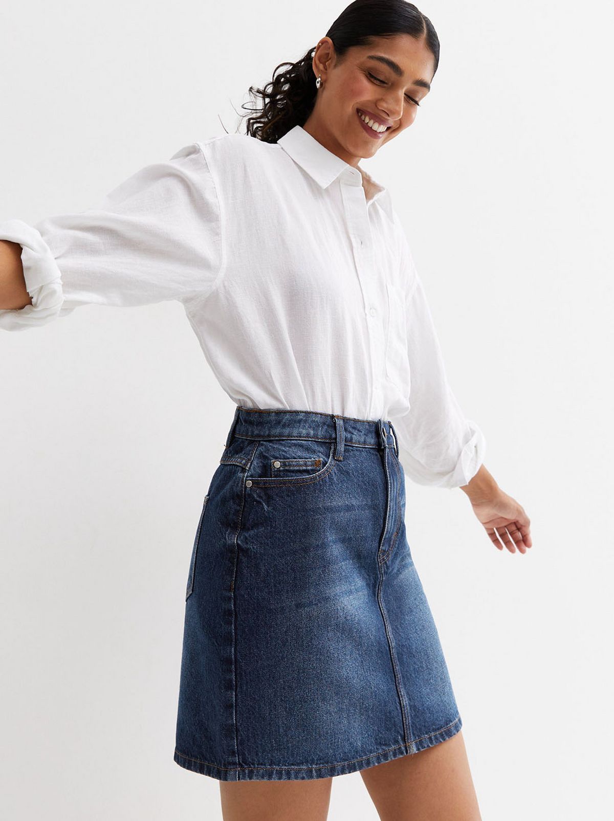 A model is standing in front of a white background while wearing a blue denim skirt and a white collar shirt	