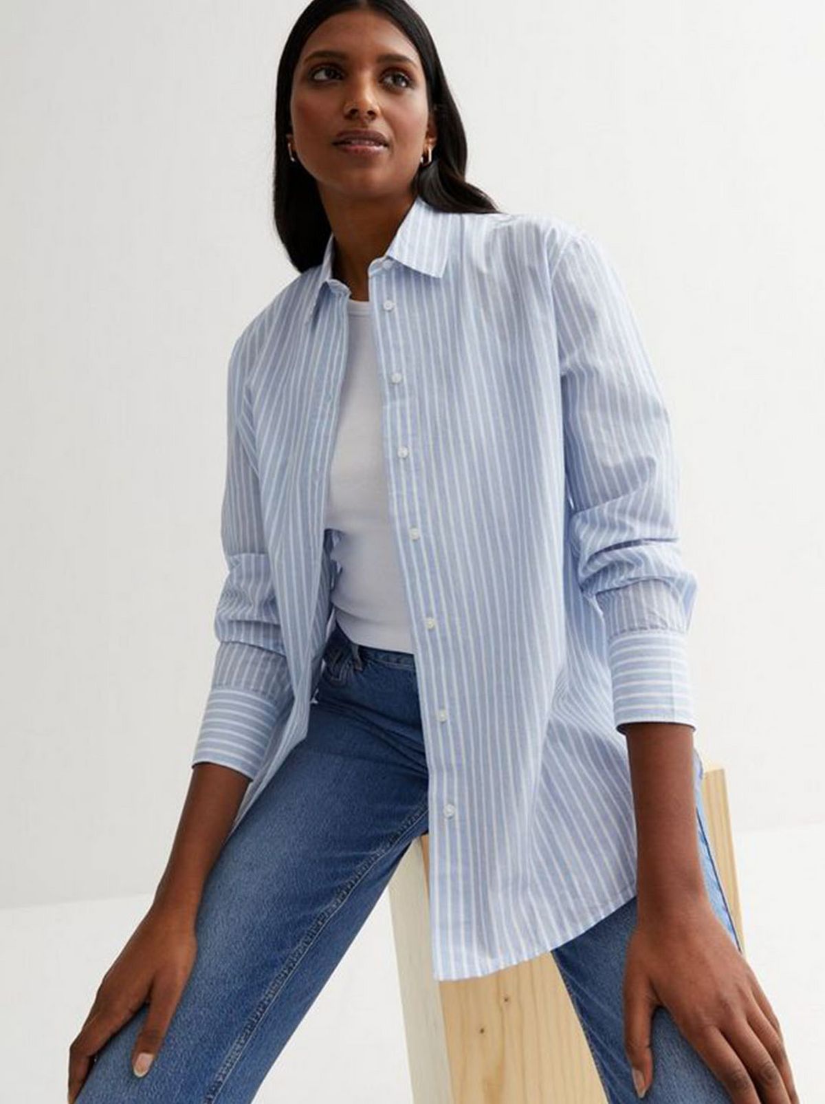 A model is sitting in front of a white background while wearing a white t shirt, a blue cotton poplin shirt and a pair of blue jeans	