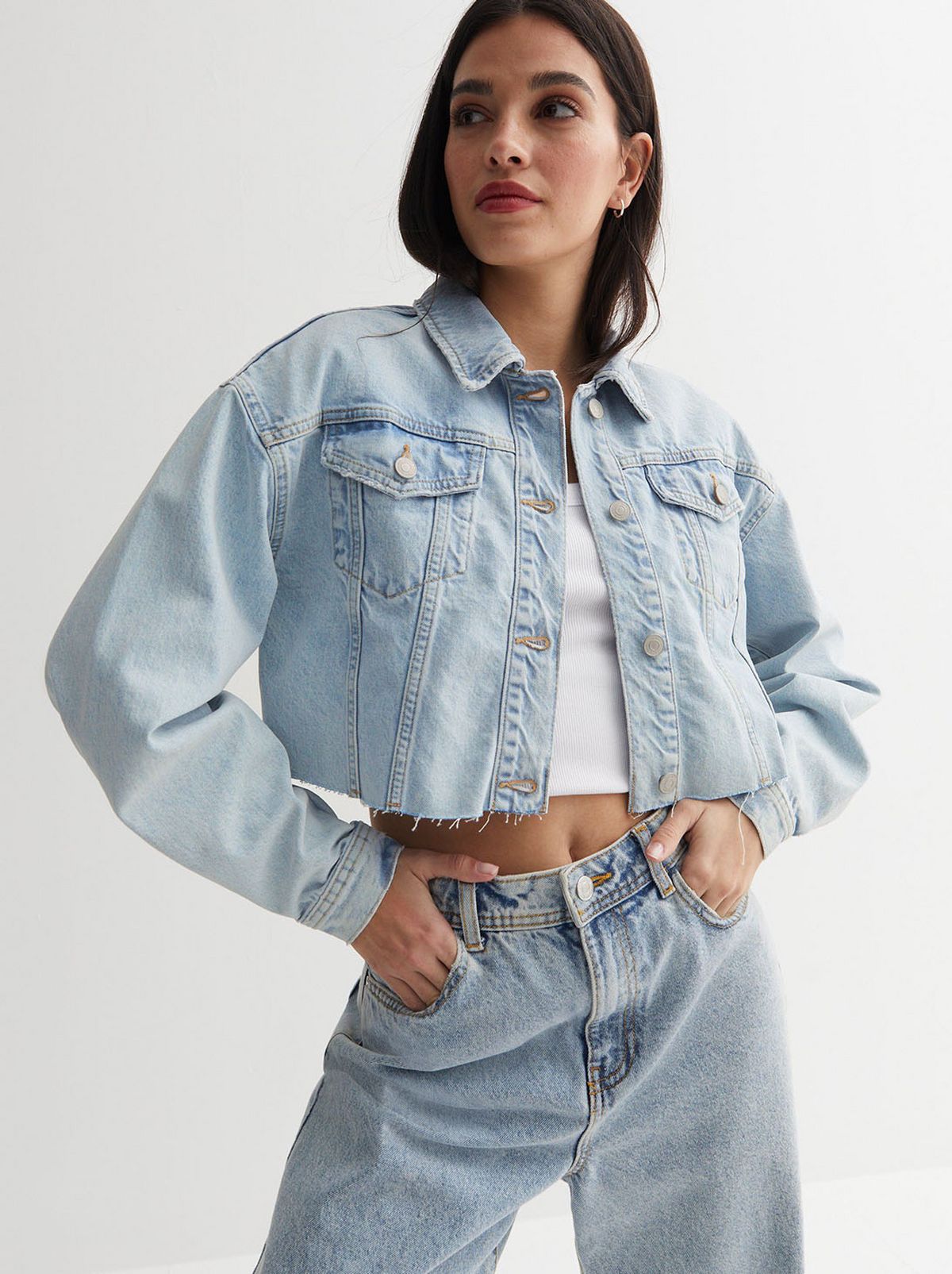 A model is standing in front of a white background while wearing a pale blue cropped denim jacket and a matching pair of jeans	