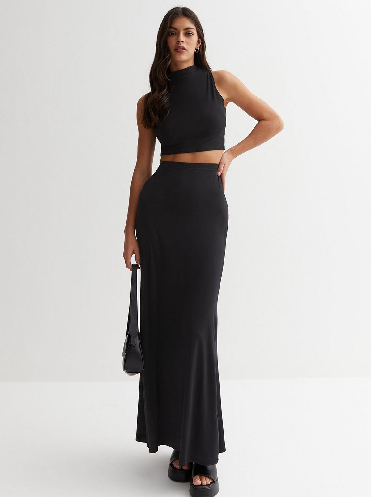 A model is standing in front of a white background while wearing a black high waist maxi skirt and a vest top	