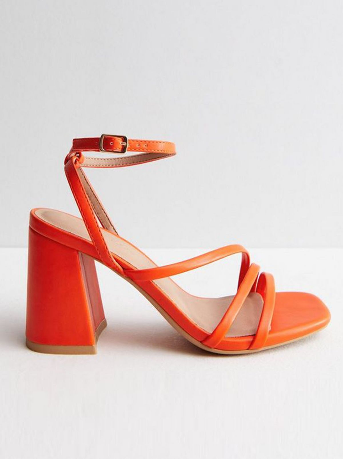A bright orange block heel sandal stands on a white floor in front of a white background