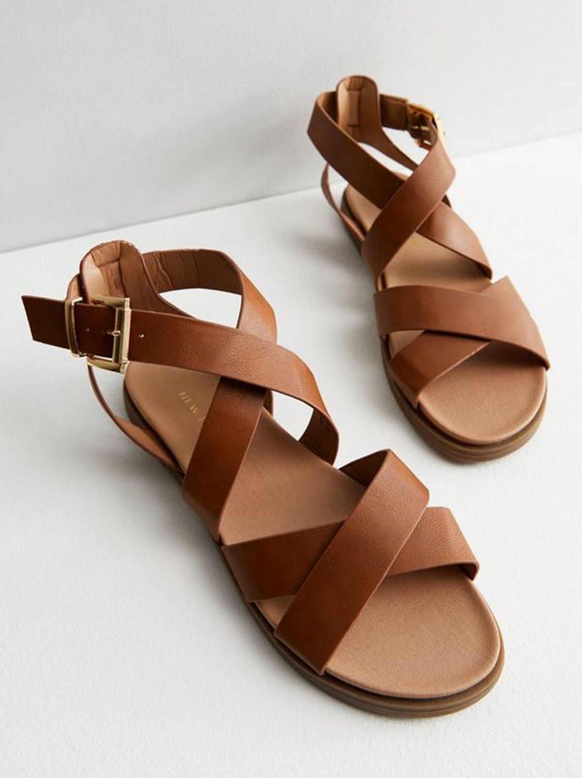A tan leather look strappy sandal stands on a white floor in front of a white background