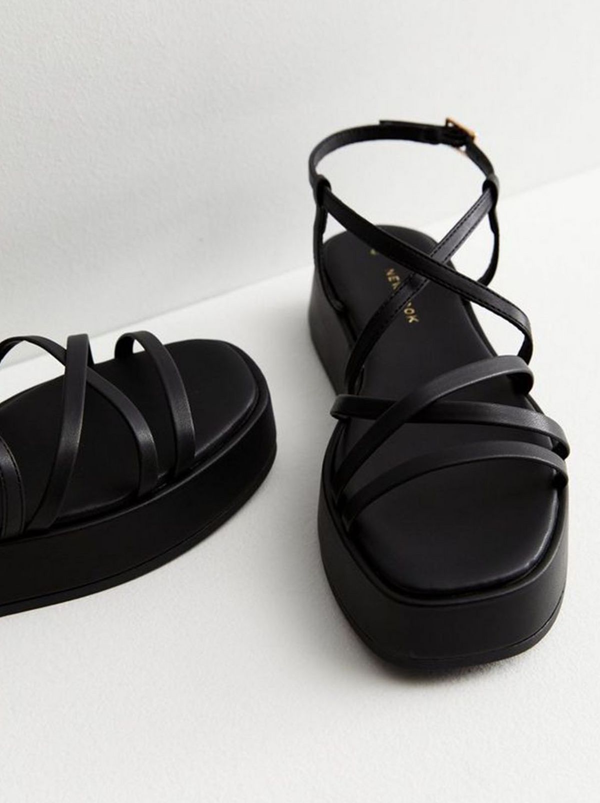 A black leather look strappy flatform sandal stands on a white floor in front of a white background