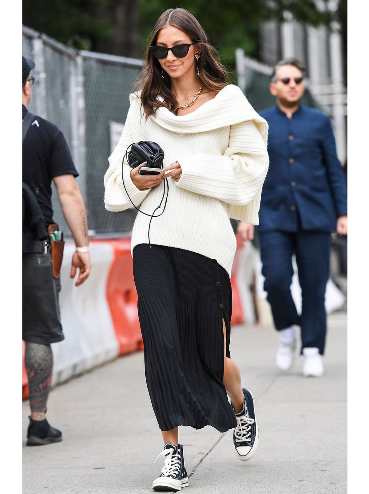 Woman walking in the street wearing a long black skirt and chunky knit jumper with sunglasses