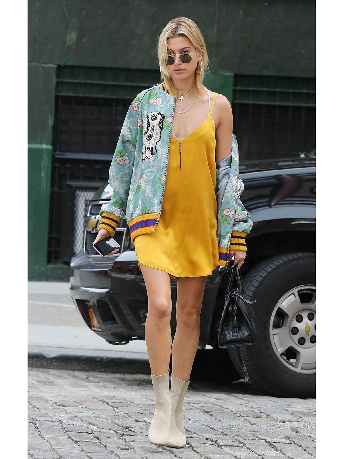 Hailey Bieber crossing the road wearing a yellow mini slip dress and oversized bomber jacket and boots